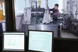 Technician operating industrial machinery in a laboratory-like facility, viewed from behind computer monitors displaying technical data.
